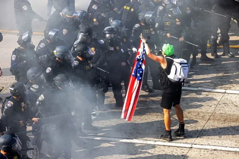 محتج يحمل علم الولايات المتحدة وسط دخان الغاز المسيل للدموع، يواجه صفوفًا من الشرطة المدججة بالسلاح في لوس أنجلوس.