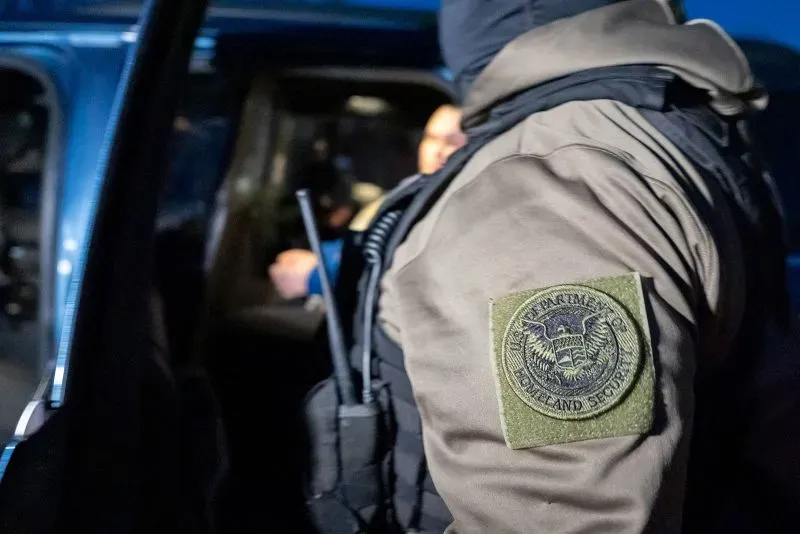 صورة تبرز موظفًا من وكالة إنفاذ قوانين الهجرة والجمارك (ICE) يرتدي زيًا رسميًا، مع شعار الوكالة على ذراعه، خلال تنفيذ عمليات إنفاذ.