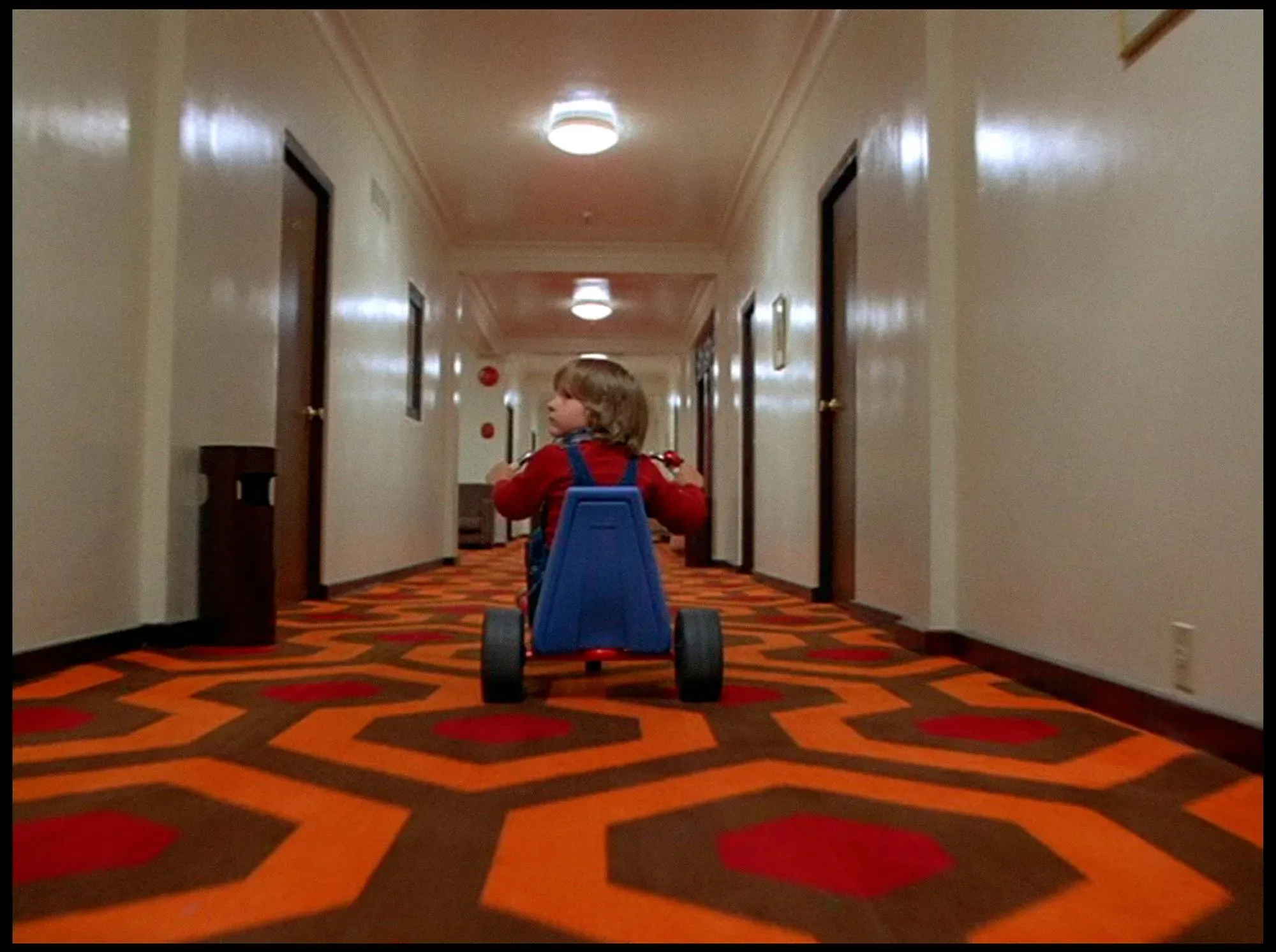 طفل يقود عربة صغيرة في ممر فندق مزين بسجاد ملون، مشهد مأخوذ من فيلم "The Shining"، يعكس أجواء الرعب والغموض.