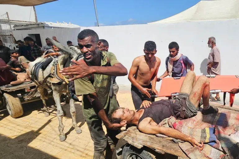 رجال يحملون مصابًا على عربة، في مركز توزيع المساعدات بغزة، وسط مشاهد من الألم والفوضى بسبب نقص الغذاء.