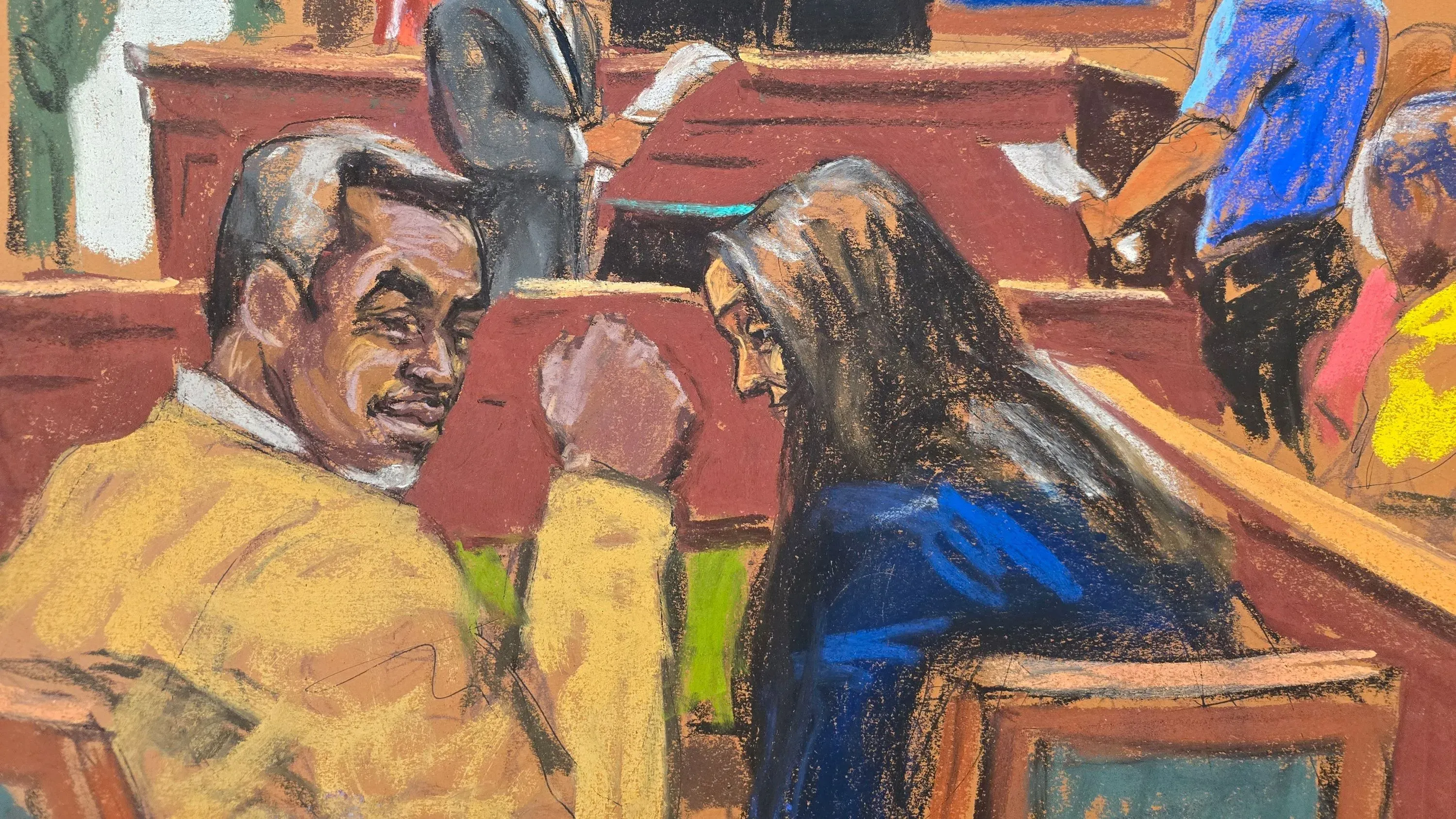 كومبس يجلس في قاعة المحكمة، مبتسمًا، بجوار امرأة ذات شعر طويل، بينما يتحدث محامو الدفاع خلفهم.