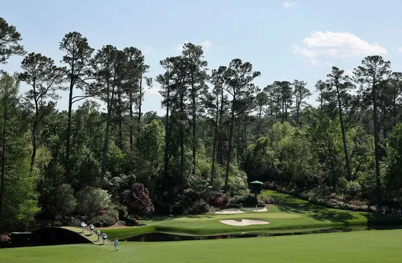 منظر لملعب الجولف في بطولة الماسترز، يظهر اللاعبين يتجهون نحو الحفرة الأولى، محاطين بالأشجار الخضراء والزهور.