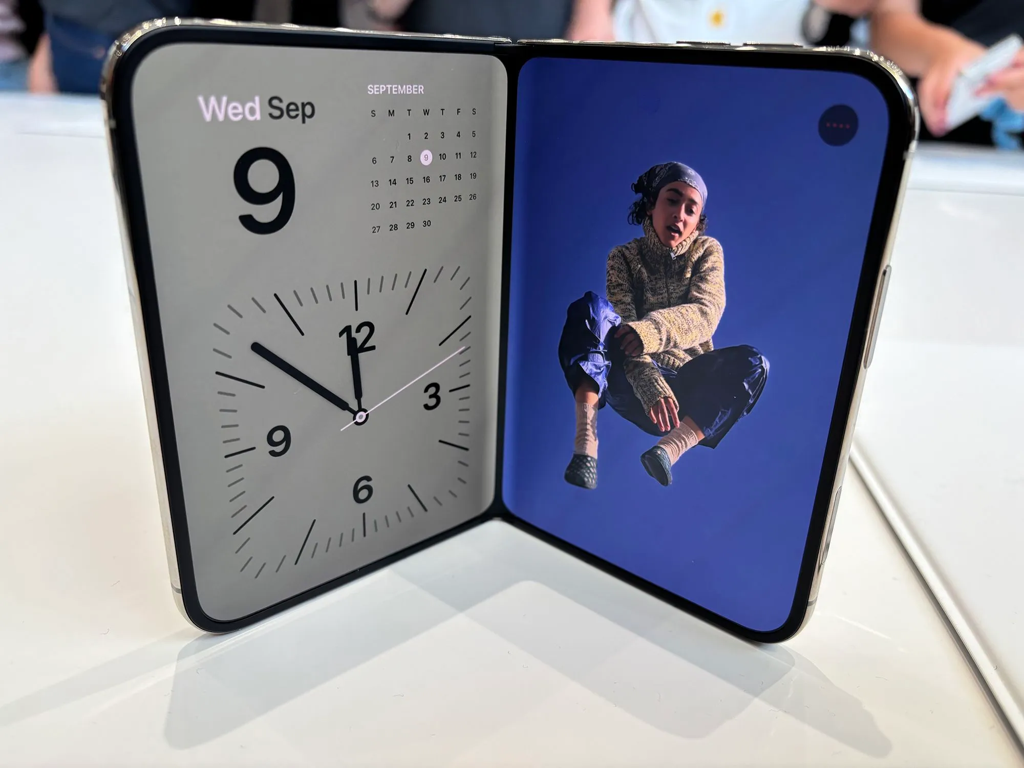 هاتف iPhone Duo القابل للطي مفتوح يعرض شاشة مزدوجة، أحدهما ساعة وتقويم والآخر صورة شخص، مع تصميم أنيق وابتكار Apple في الهواتف الذكية.