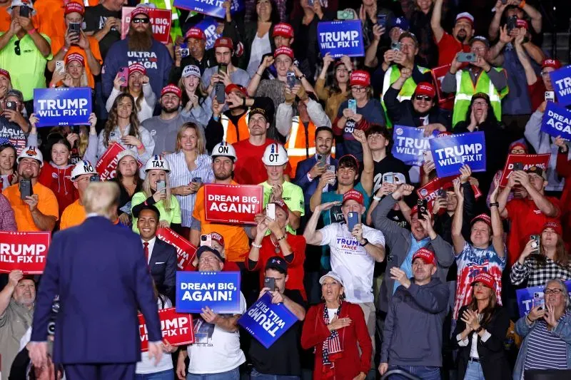 حشود من مؤيدي ترامب يرتدون قبعات وقمصان تحمل شعارات انتخابية، يرفعون لافتات تدعم حملته خلال تجمع انتخابي.