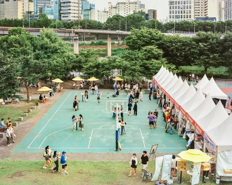 مشهد حيوي لمهرجان في حديقة بمدينة سيول، حيث يتجمع الناس حول ملعب كرة السلة، تحت الأشجار، مع أكشاك تعرض المنتجات.