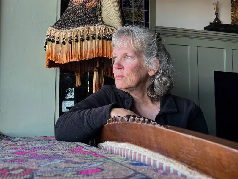 امرأة مسنّة تجلس بجانب نافذة، تنظر بتفكير إلى المسافة، مع وجود مصباح مزخرف خلفها، تعكس مشاعر القلق إثر حرائق الغابات في كولورادو.