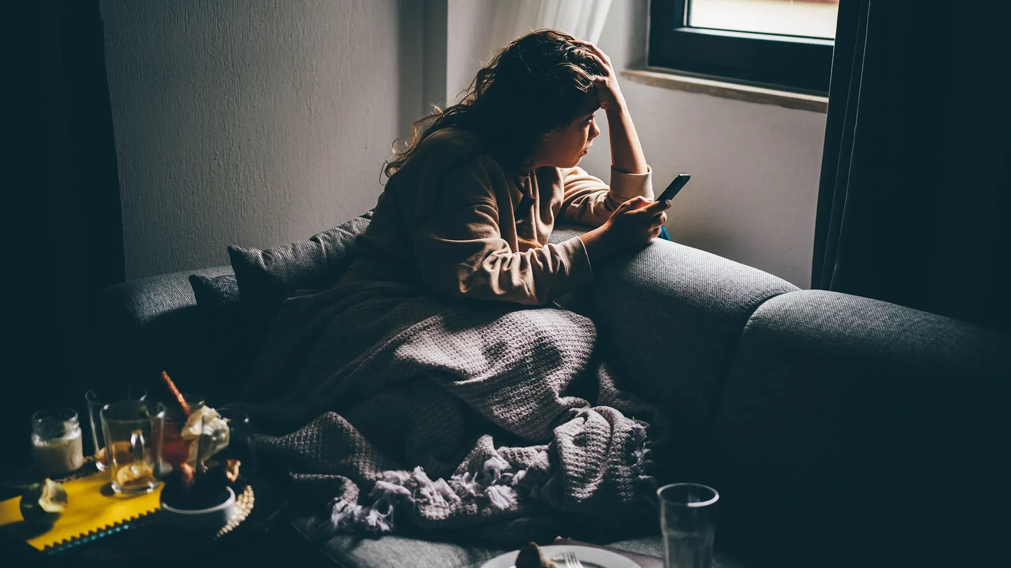 امرأة جالسة على أريكة، تمسك بهاتفها، تبدو مشغولة بالتفكير، محاطة بأشياء تشير إلى العزلة والتأمل في تأثير وسائل التواصل الاجتماعي على حياتها.