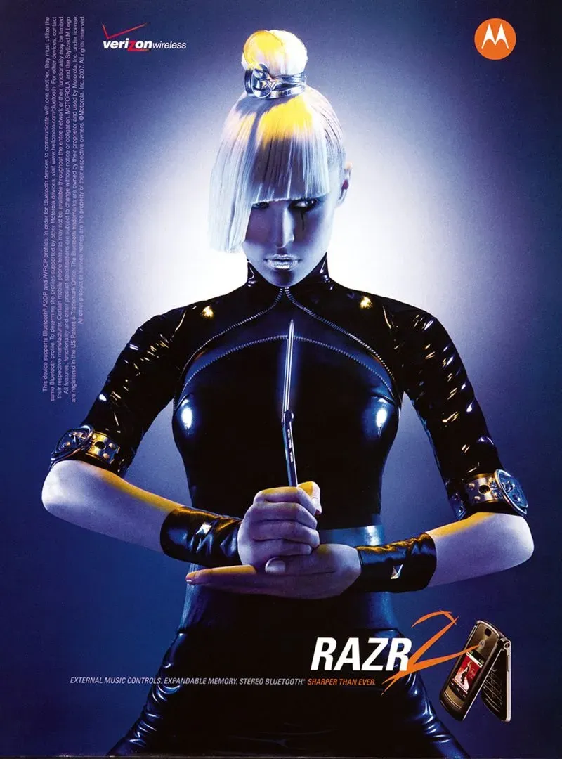 امرأة ترتدي بدلة جلدية سوداء، تحمل سكينًا، في إعلان لهاتف Motorola RAZR2، مع إضاءة خلفية زرقاء تعزز الأجواء المستقبلية.