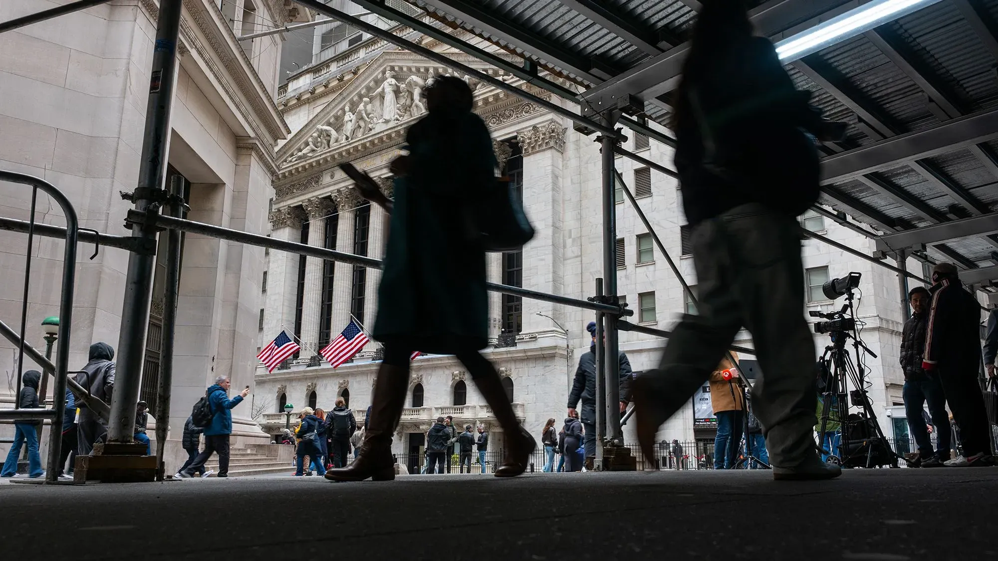 مشهد من أمام بورصة نيويورك، حيث يسير الناس تحت هيكل مؤقت، مع أعلام أمريكا ترفرف في الخلفية، مما يعكس حالة السوق المالية.