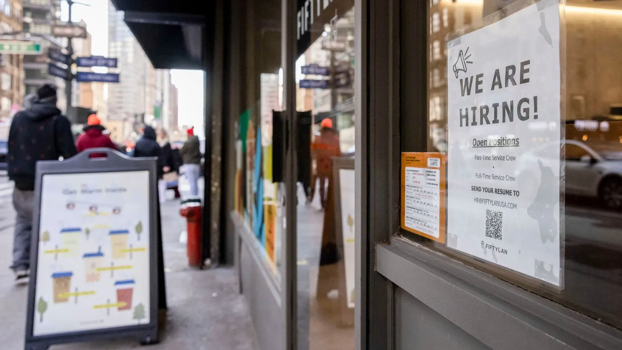 لافتة "نحن نوظف!" معلقة على واجهة متجر، تظهر فرص العمل المتاحة في الخدمة الجزئية والكاملة، مع وجود أشخاص يمشون في الشارع.
