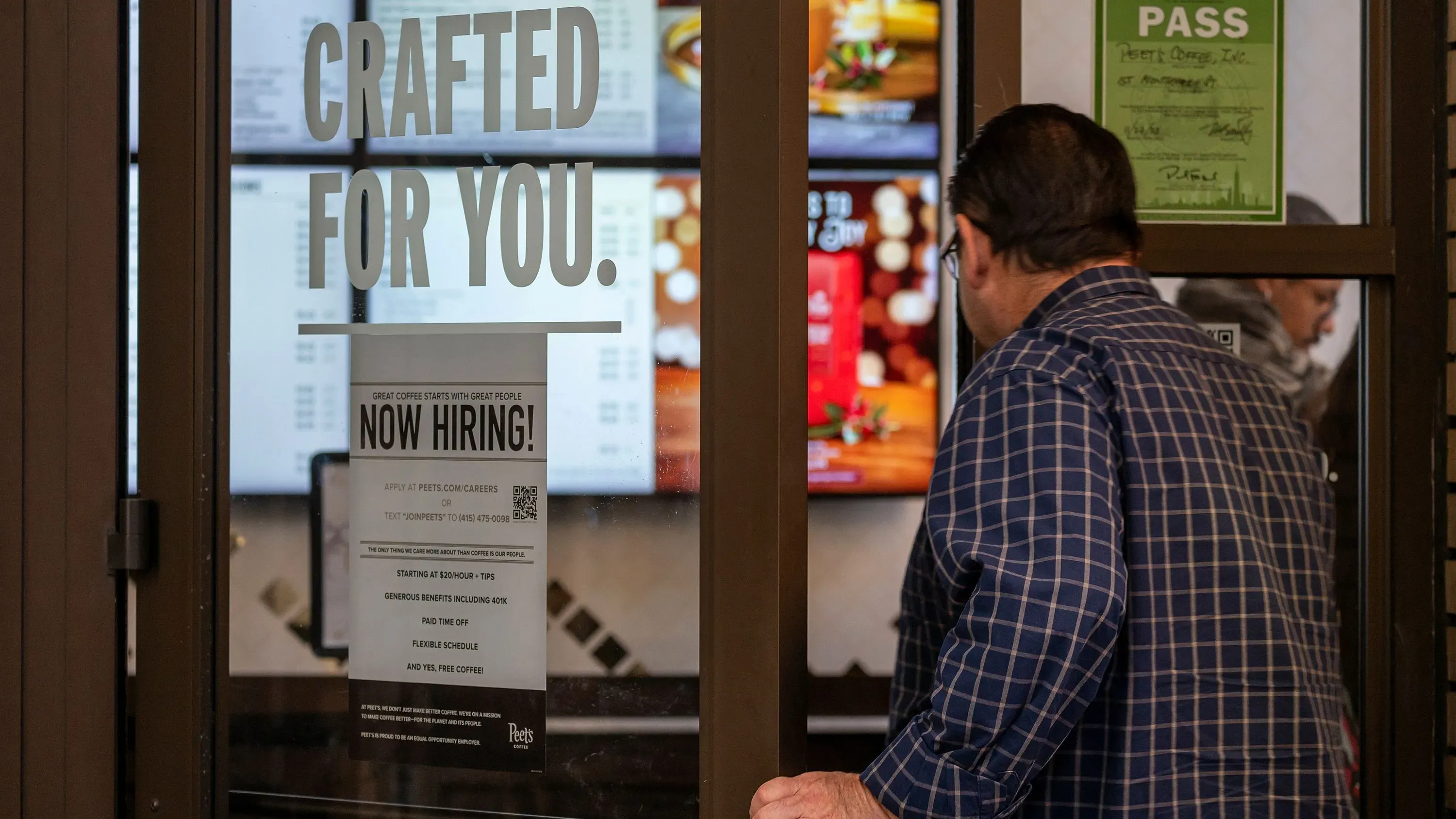 رجل يدخل مقهى يحمل لافتة "نحن نوظف!"، مما يعكس حالة سوق العمل في ظل التغيرات الاقتصادية الحالية.