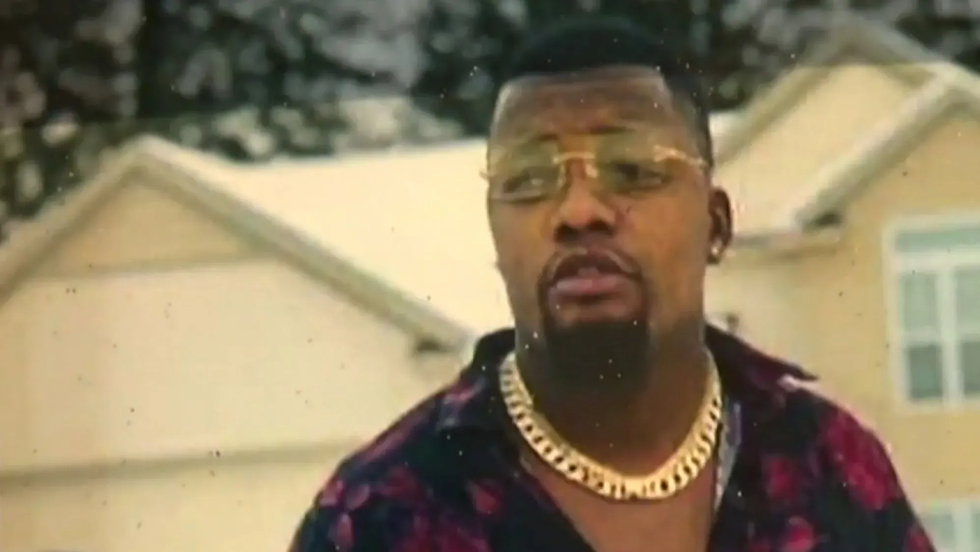 صورة لرجل يرتدي نظارات شمسية وسلسلة ذهبية، يتحدث في مكان مفتوح، مع خلفية منزلية. تعكس الصورة القضايا الاجتماعية المحيطة بمقتل باتريك ليويا.