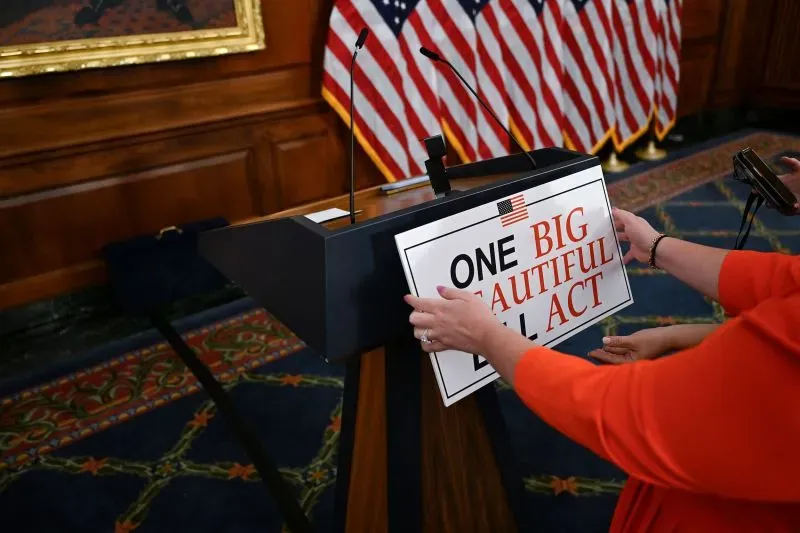 شخصية ترتدي سترة برتقالية تضع لافتة مكتوب عليها "مشروع قانون واحد كبير وجميل" على منصة في قاعة مجلس الشيوخ، مع العلم الأمريكي في الخلفية.
