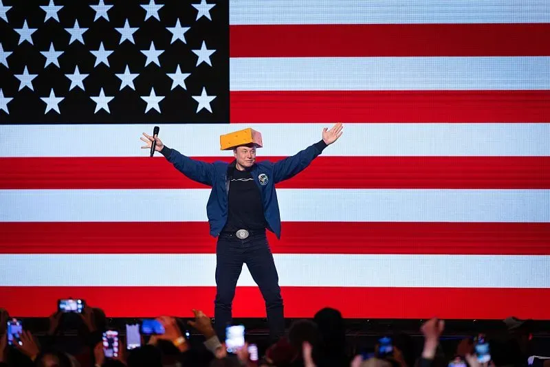إيلون ماسك يرتدي قبعة من الجبنة في حدث سياسي، مع العلم الأمريكي في الخلفية، مما يعكس تأثيره في الانتخابات بولاية ويسكونسن.
