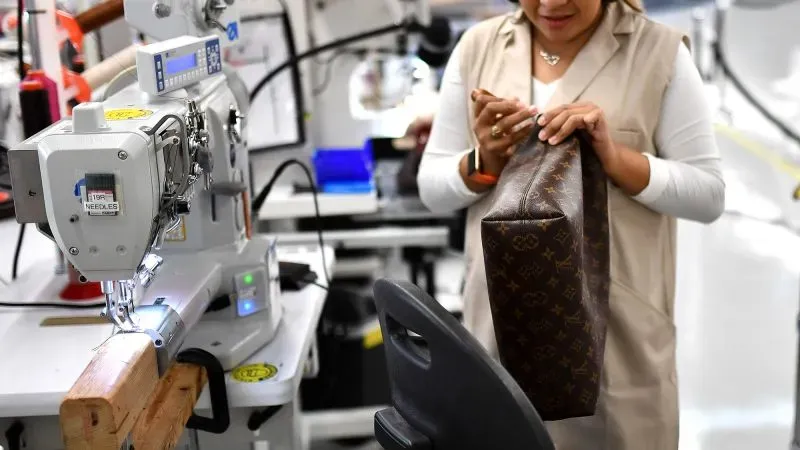 امرأة تعمل في مصنع للأزياء، تمسك بحقيبة فاخرة أثناء استخدام آلة خياطة. تعكس الصورة تأثير التعريفات الجمركية على صناعة الملابس.