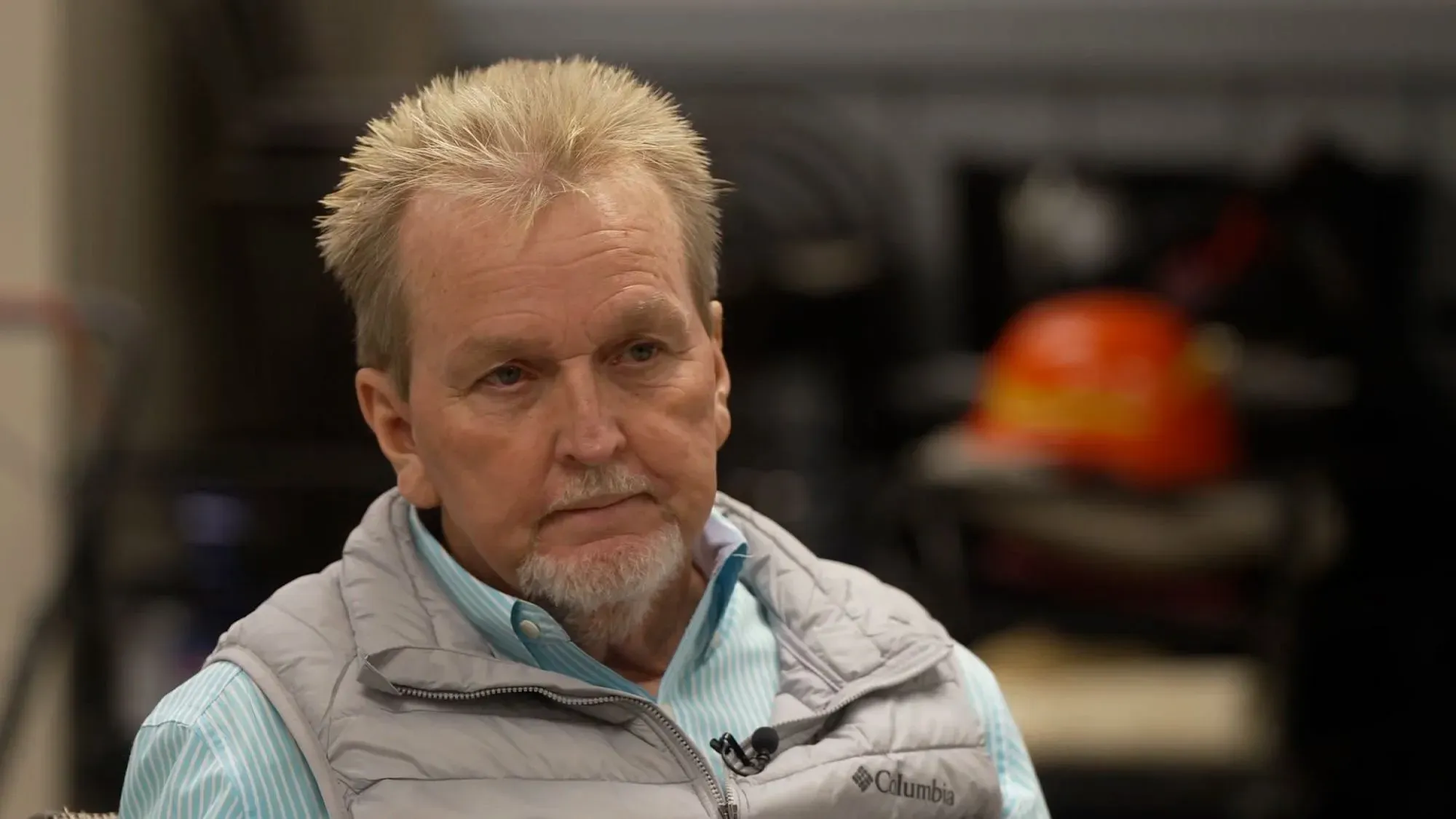 رجل مسن ذو لحية قصيرة يجلس في مكان داخلي، يبدو عليه التفكير العميق، مع أدوات خلفه تشير إلى بيئة عمل.