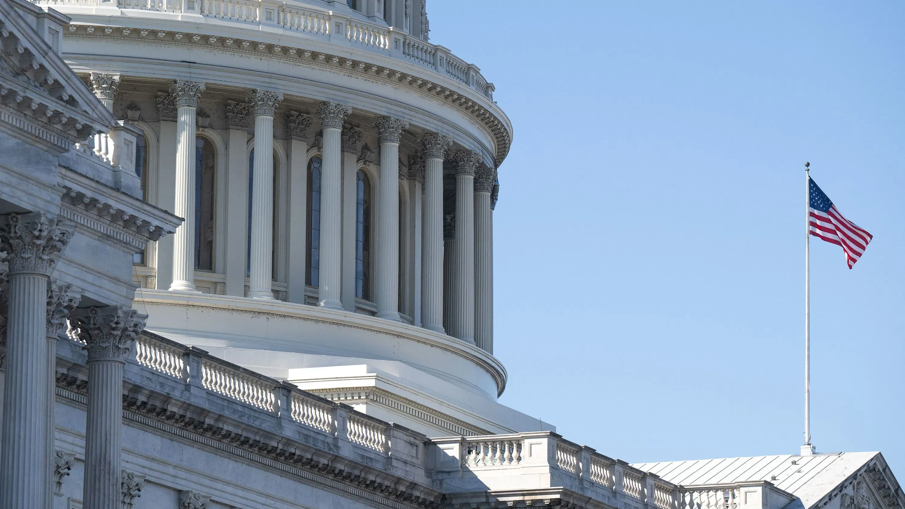 صورة لجزء من مبنى الكابيتول الأمريكي يظهر أعمدة معمارية مميزة وعلم الولايات المتحدة يرفرف في السماء الزرقاء.