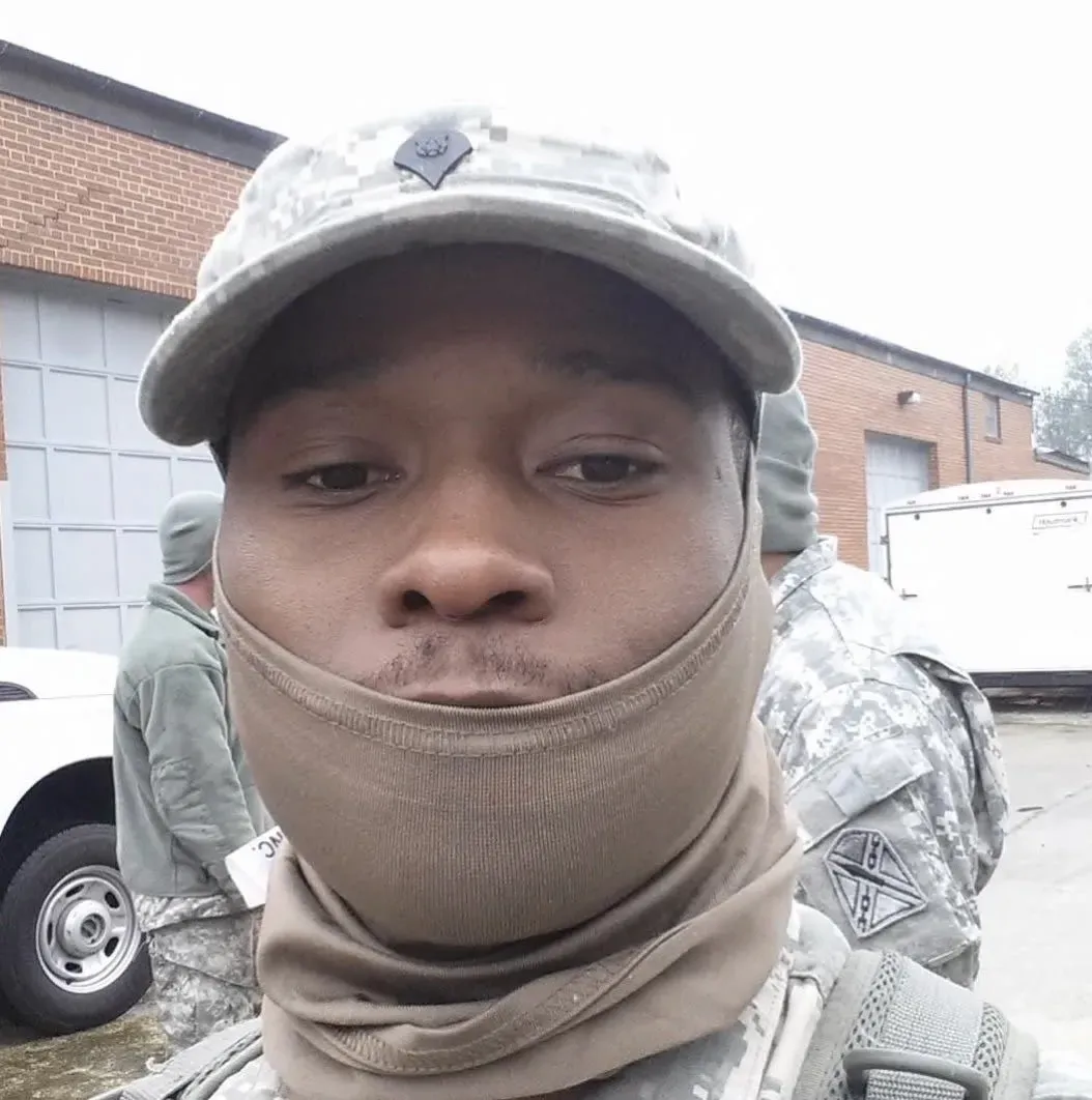 صورة لمسلح سابق في الحرس الوطني الأمريكي، محمد بيلور جالوه، يرتدي زيًا عسكريًا وقبعة، مع خلفية تظهر أشخاصًا آخرين في ملابس عسكرية.