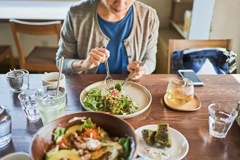 امرأة تتناول سلطة صحية في مطعم، مع أطباق متنوعة على الطاولة، تعكس أهمية التغذية السليمة للشيخوخة الصحية.