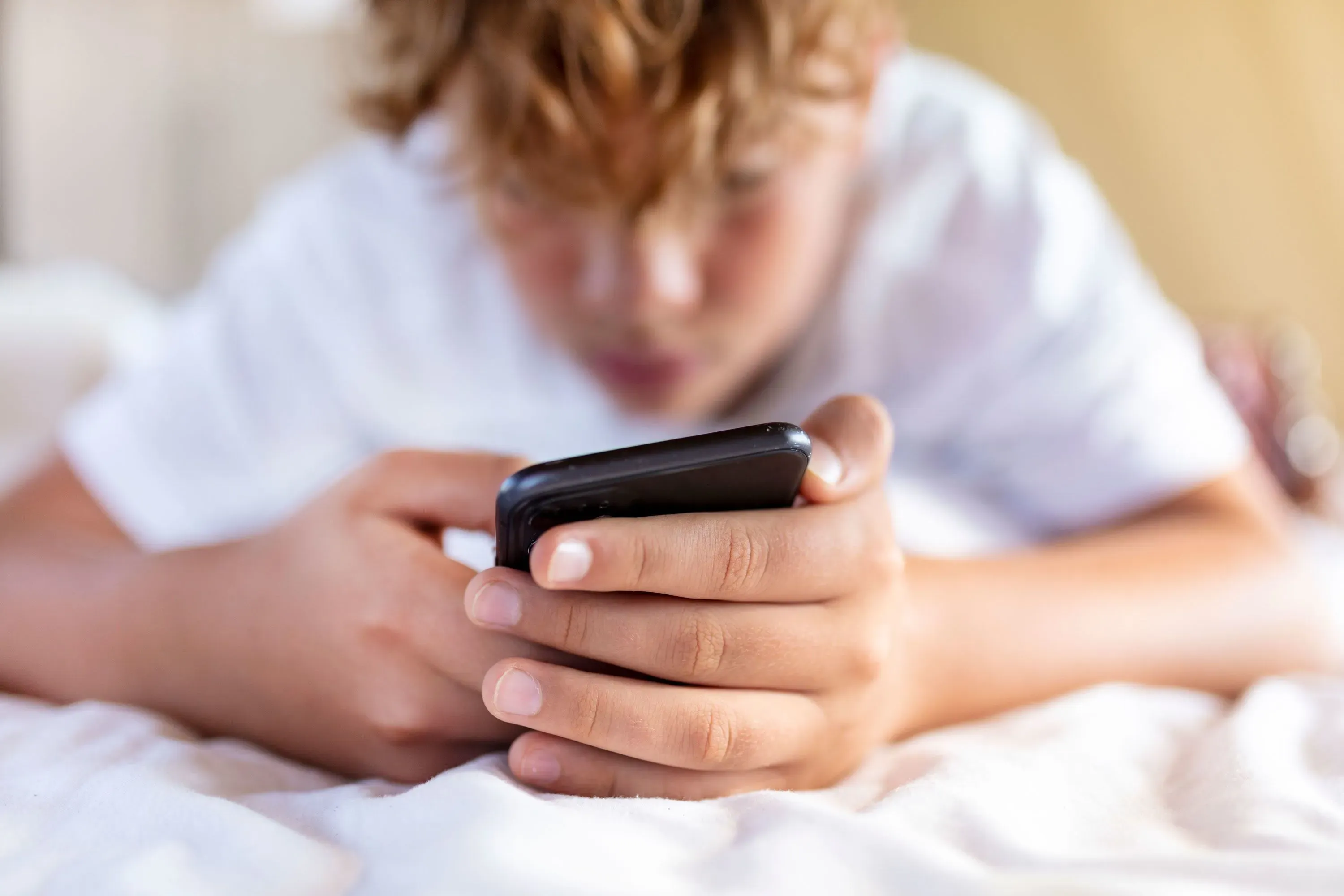 صبي مراهق يجلس على السرير، ينظر إلى هاتفه الذكي بتركيز. تعكس الصورة تأثير وسائل التواصل الاجتماعي على الشباب.