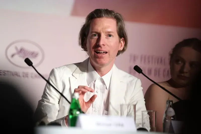 مؤتمر صحفي في مهرجان كان السينمائي، حيث يتحدث رجل يرتدي بدلة بيضاء، مع زجاجة مياه على الطاولة خلفه، بينما تظهر امرأة في الخلفية.