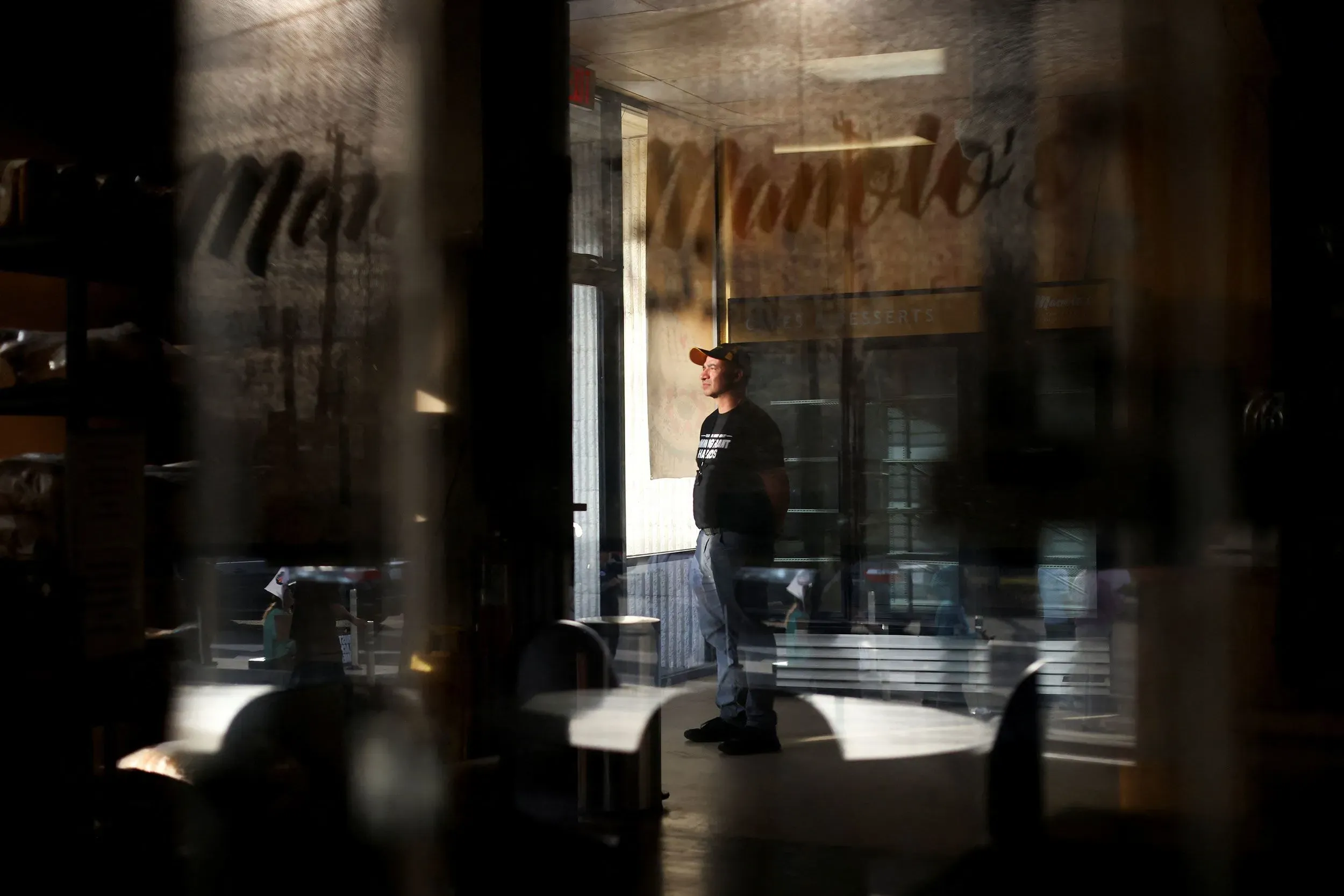 رجل يقف داخل مطعم، يتأمل من خلال نافذة زجاجية، بينما تعكس الإضاءة المخاوف من مداهمات الهجرة في مجتمعه.