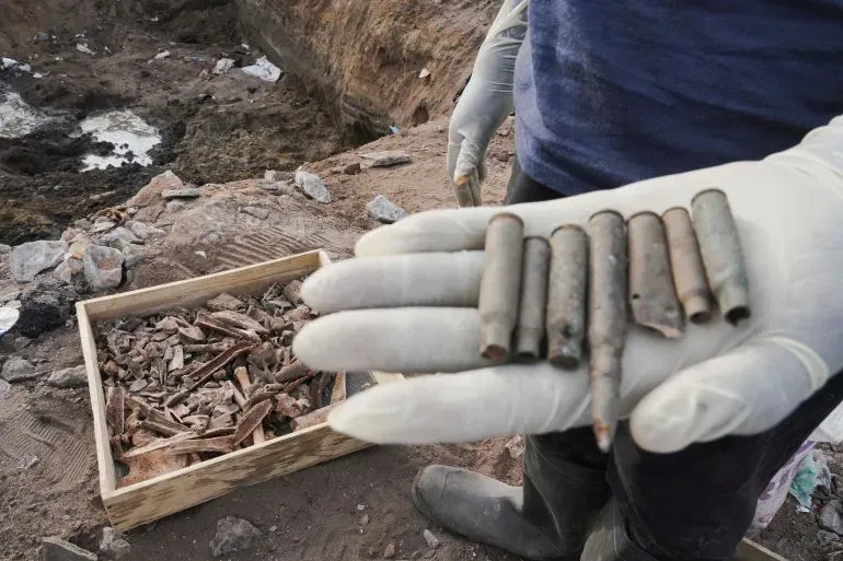 عالم آثار يحمل مجموعة من الطلقات الفارغة في يده، مع وجود حفر خلفه تحتوي على بقايا وأدوات، مما يشير إلى نشاط استكشافي.