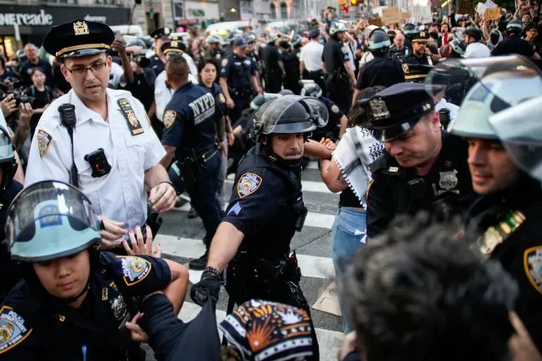 اشتباكات بين الشرطة ومحتجين خلال مظاهرة ضد مداهمات الهجرة في مدينة مزدحمة، مع وجود عناصر من الشرطة يرتدون خوذات واقية.