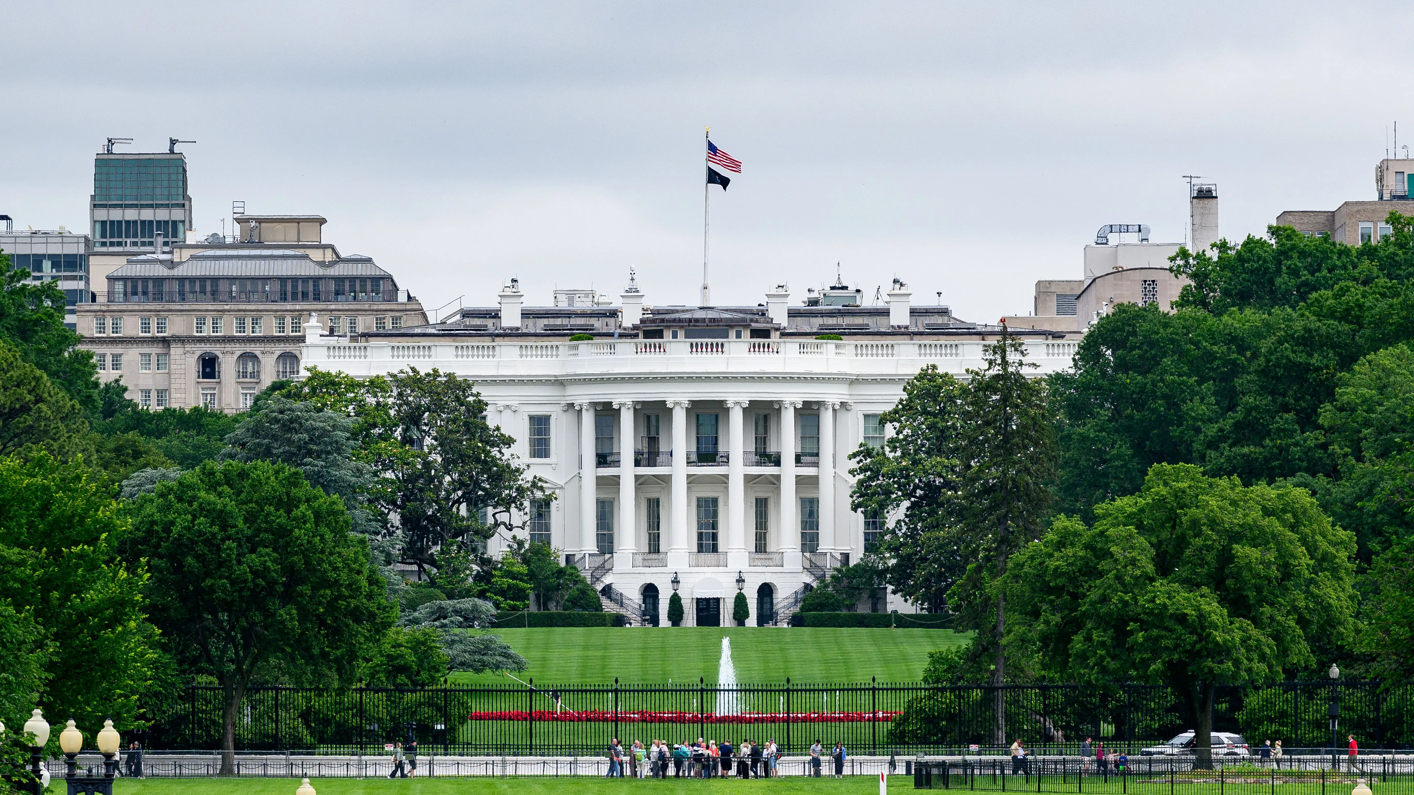 البيت الأبيض يظهر في صورة تحت سماء غائمة، مع العلم الأمريكي يرفرف فوقه، بينما يتجمع عدد من الأشخاص في الحديقة.