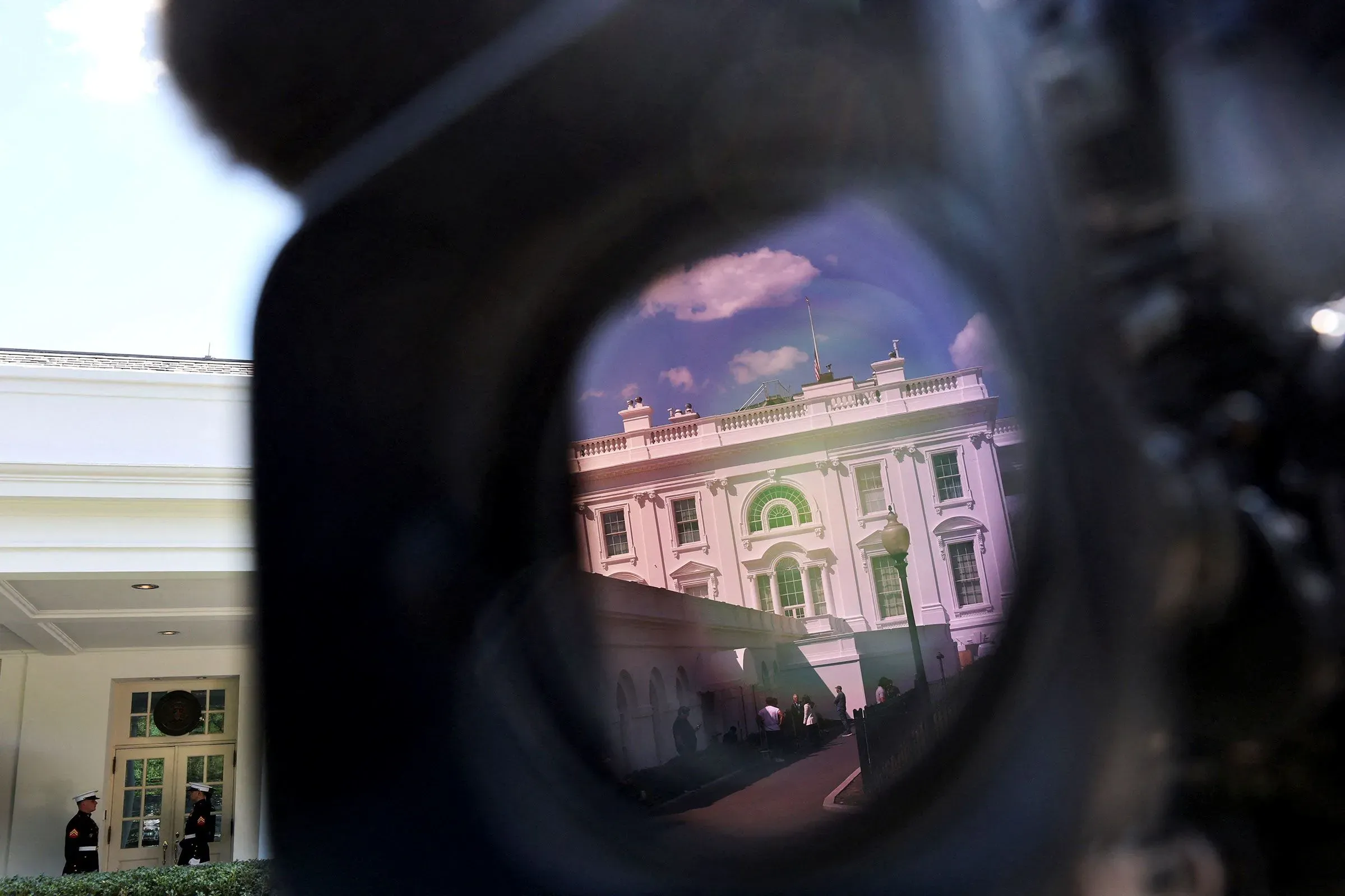 من خلال عدسة كاميرا، يظهر البيت الأبيض مع سماء زرقاء وصافية، بينما يتواجد حراس أمام المدخل. يعكس المشهد التوترات السياسية الجارية.