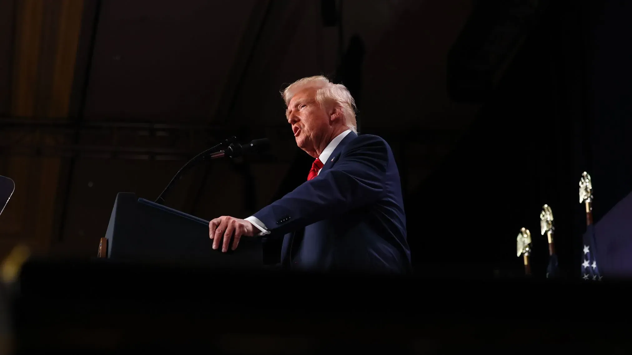 تظهر الصورة الرئيس السابق ترامب أثناء إلقائه خطابًا، حيث يتحدث إلى الجمهور وسط أجواء رسمية، مع وجود علم خلفه.