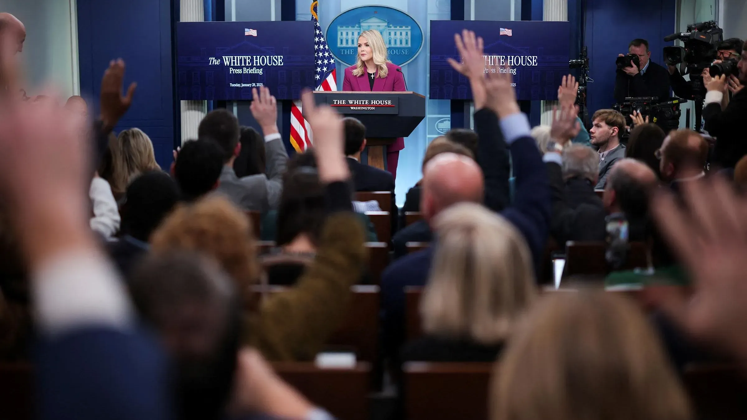 إحاطة صحفية في البيت الأبيض حيث تتحدث كارولين ليفيت وسط جمهور يرفع أيديه لطرح الأسئلة، مع وجود شاشات تعرض معلومات عن المؤتمر.