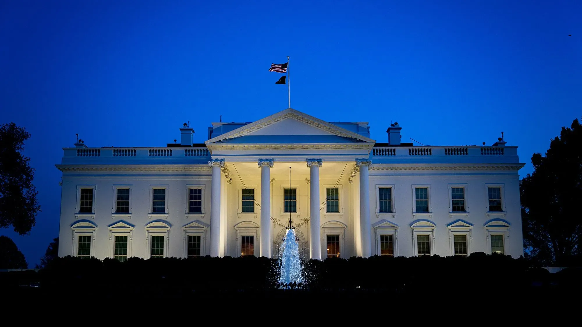البيت الأبيض مضاء في المساء، مع العلم الأمريكي يرفرف فوقه، مما يعكس الجدل حول أصول فيروس كورونا والتسريبات المخبري.