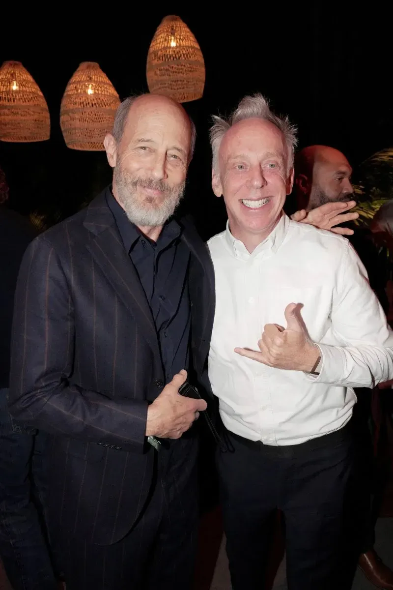 صورة لجون جريس ومبدع مسلسل "اللوتس الأبيض" مايك وايت، يتبادلان الابتسامات في حدث ترويجي، مع إضاءة خافتة وخلفية دافئة.