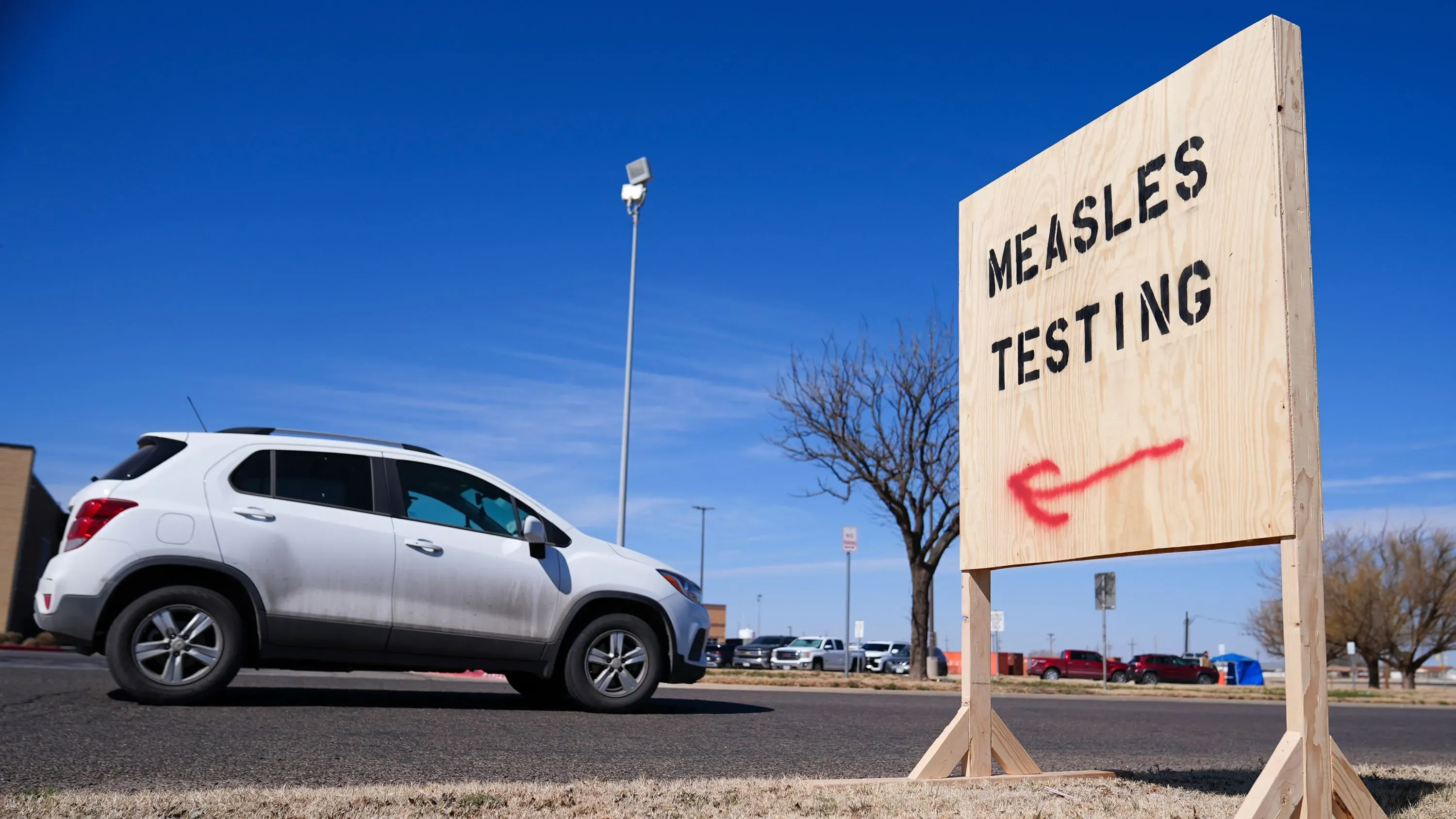 علامة خشبية تشير إلى اختبار الحصبة، مع سيارة تمر بجوارها في منطقة مفتوحة تحت سماء زرقاء.