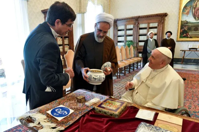 اجتماع بين رجال دين إيرانيين والبابا فرانسيس، حيث يتبادلون الهدايا في قاعة رسمية، مع وجود مكتبة في الخلفية.