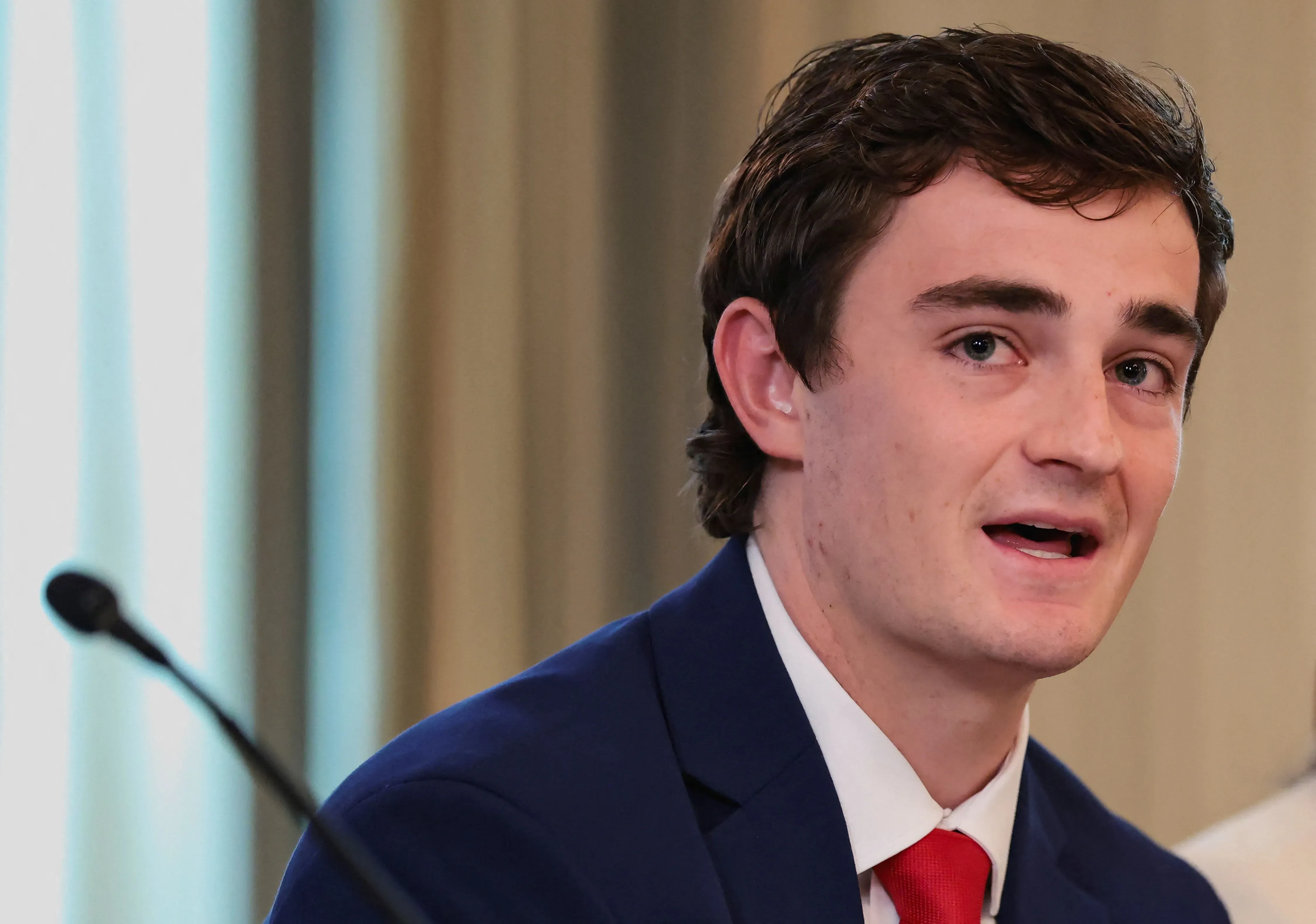 نيك شيرلي، ناشط وصحفي مستقل، يتحدث في مؤتمر حول قضايا سياسية، مرتديًا بدلة زرقاء ورابطة عنق حمراء.