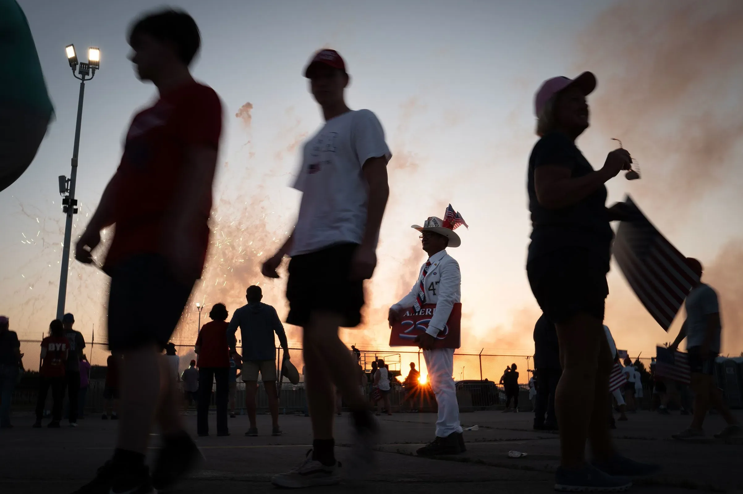 مؤيدون للرئيس ترامب في تجمع حاشد، مع شخص يرتدي زيًا مميزًا يحمل لافتة، بينما تضيء الألعاب النارية في الخلفية.