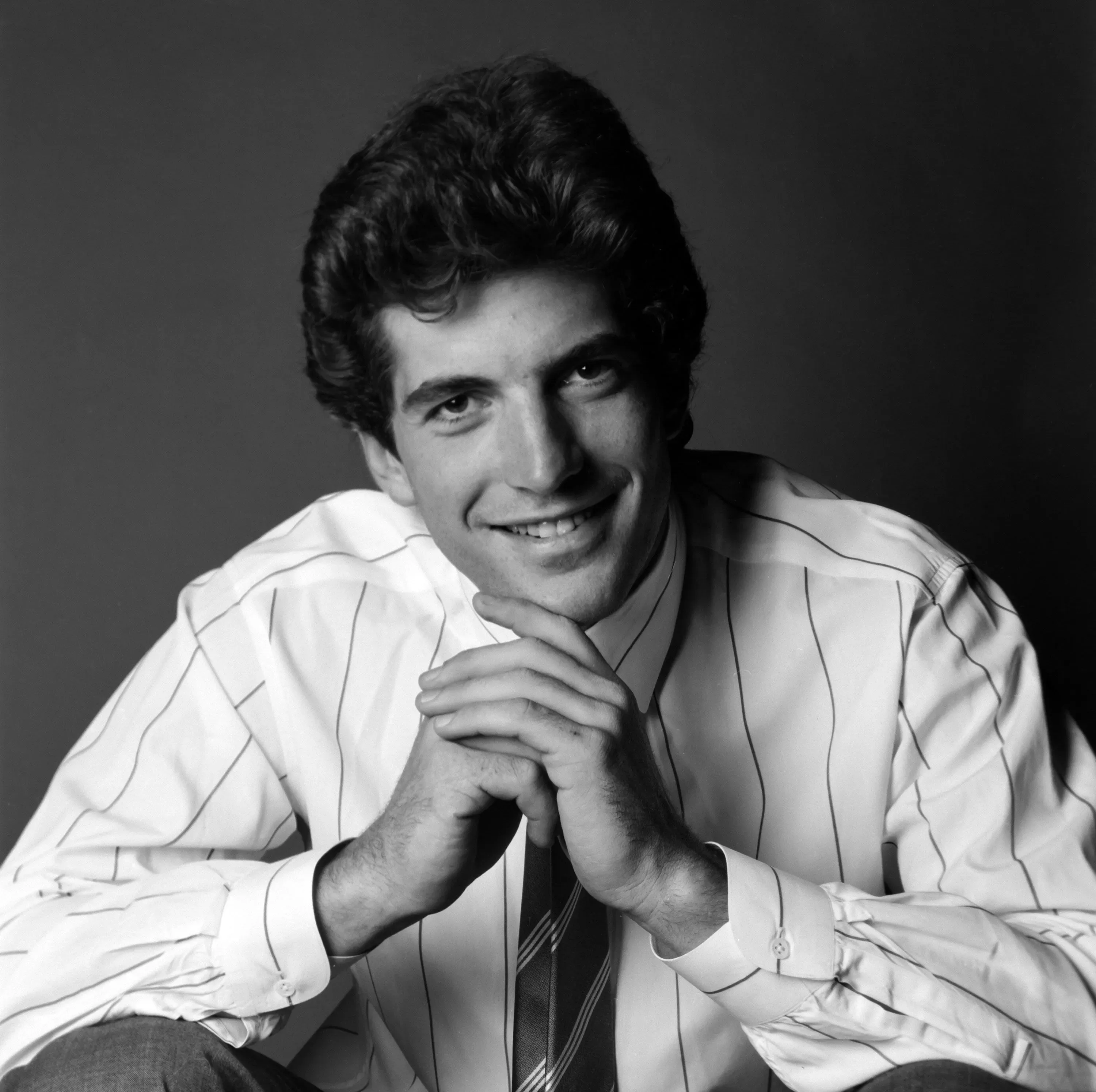 جون كينيدي الابن مبتسم في صورة بالأبيض والأسود، مرتديًا قميصًا مخططًا، مع وضع يديه تحت ذقنه، مما يعكس شخصيته الجذابة كجزء من التراث السياسي الأمريكي.