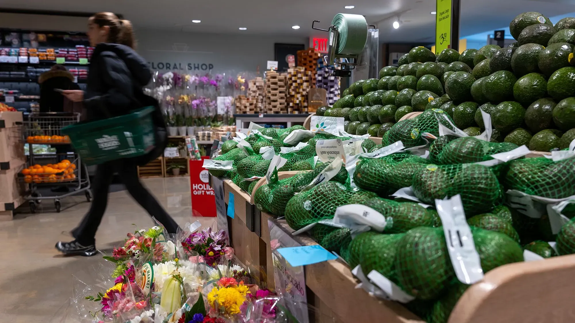 متسوقة تحمل سلة في متجر بقالة، محاطة بأفوكادو وزهور، تعكس التأثيرات المحتملة للهجوم الإلكتروني على مخزون المتاجر.