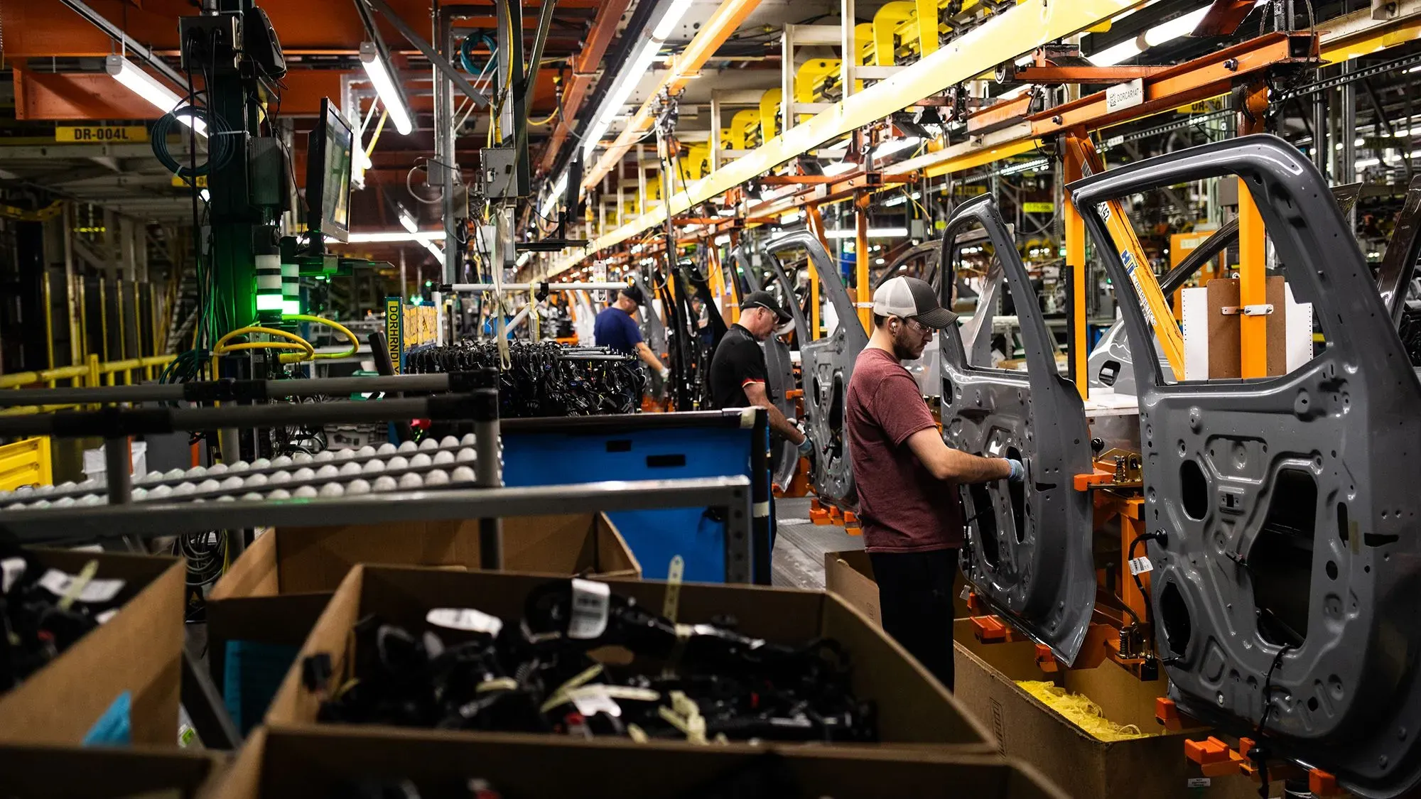عمال في مصنع يجمعون أجزاء السيارات، مع وجود معدات وأدوات في الخلفية، مما يعكس زيادة في ضغط الأسعار.