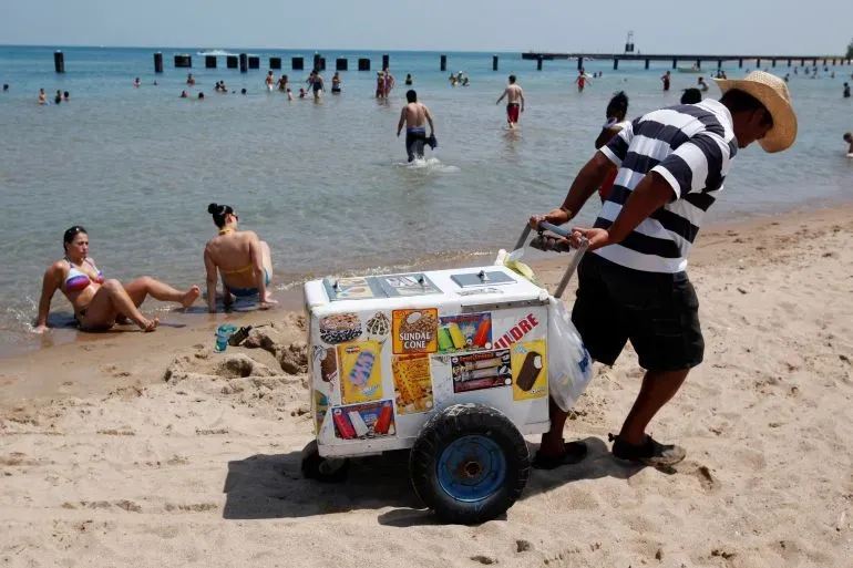 بائع آيس كريم يدفع عربة محملة بالمثلجات على شاطئ مزدحم، حيث يستمتع الناس بالسباحة في البحر.
