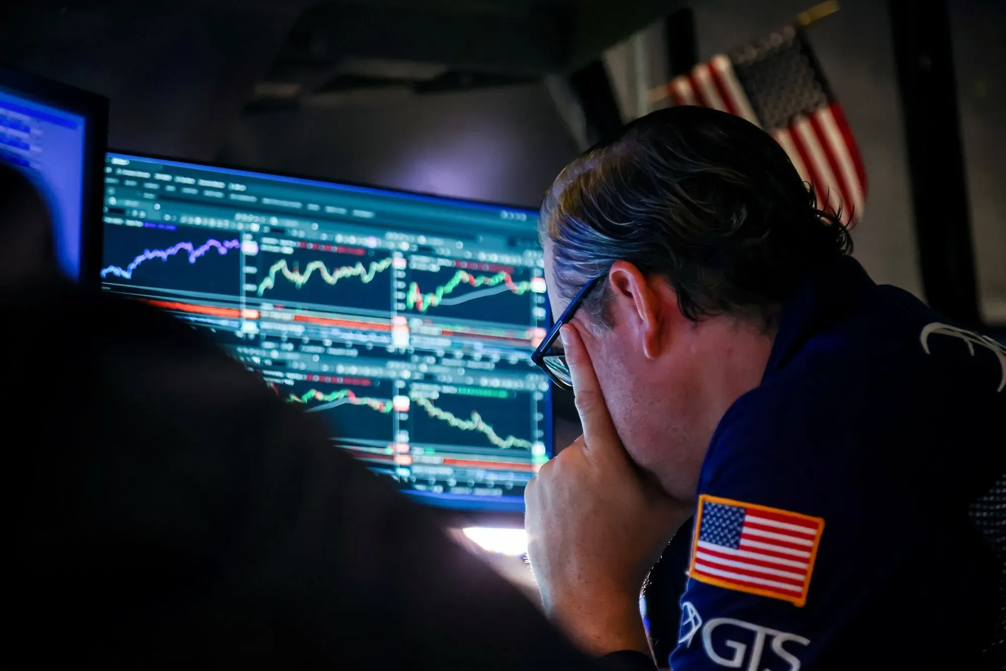 تاجر في سوق الأسهم يركز على شاشات عرض البيانات المالية، مع وجود علم الولايات المتحدة في الخلفية، مما يعكس تقلبات السوق الحالية.