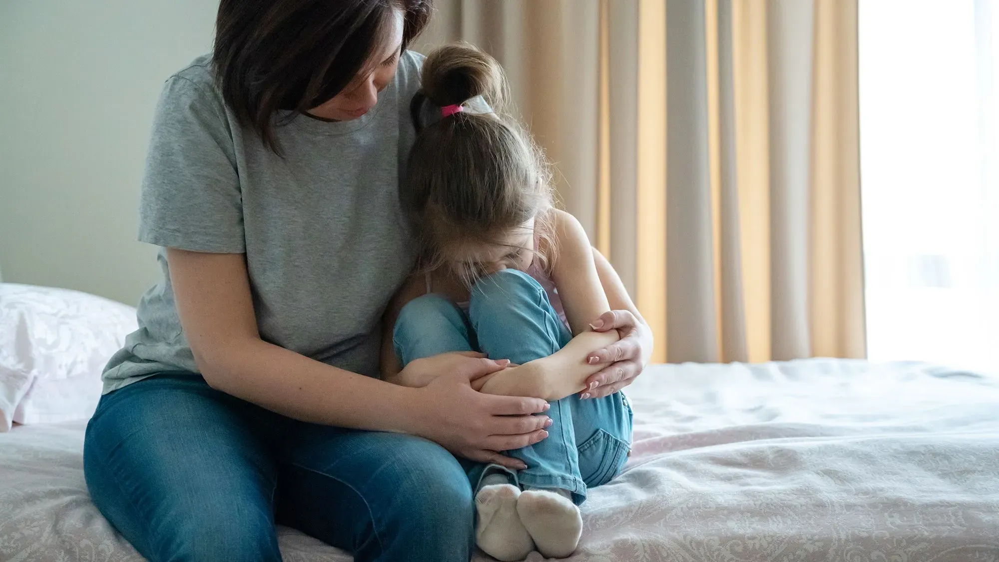 امرأة تحتضن فتاة صغيرة تجلس على السرير، تعبر عن مشاعر القلق أو الحزن. الصورة تعكس التحديات التي تواجه الفتيات المصابات بالتوحد.