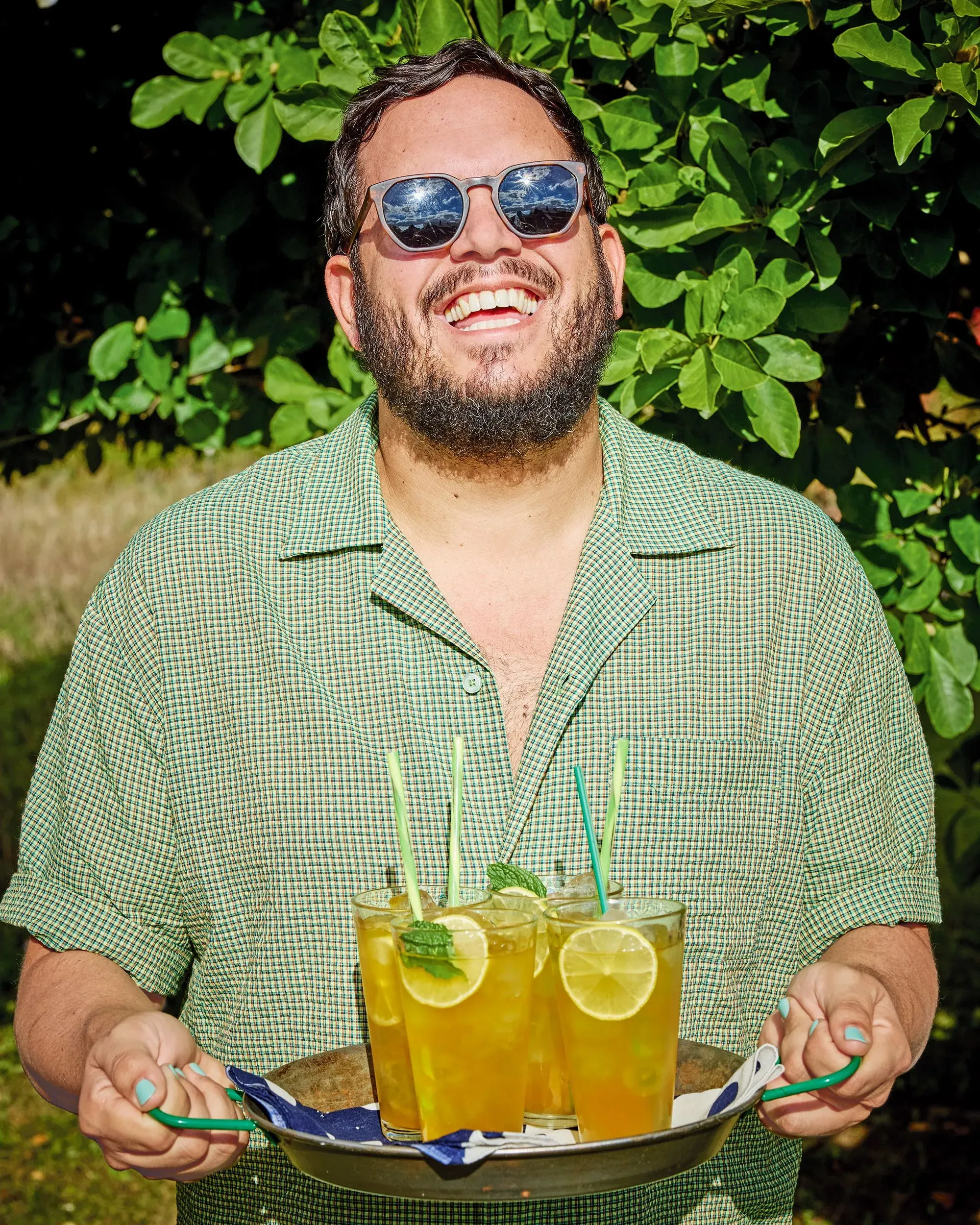 رجل مبتسم يرتدي نظارات شمسية، يحمل صينية تحتوي على مشروبات مثلجة مع شرائح الليمون، محاط بأوراق شجر خضراء.