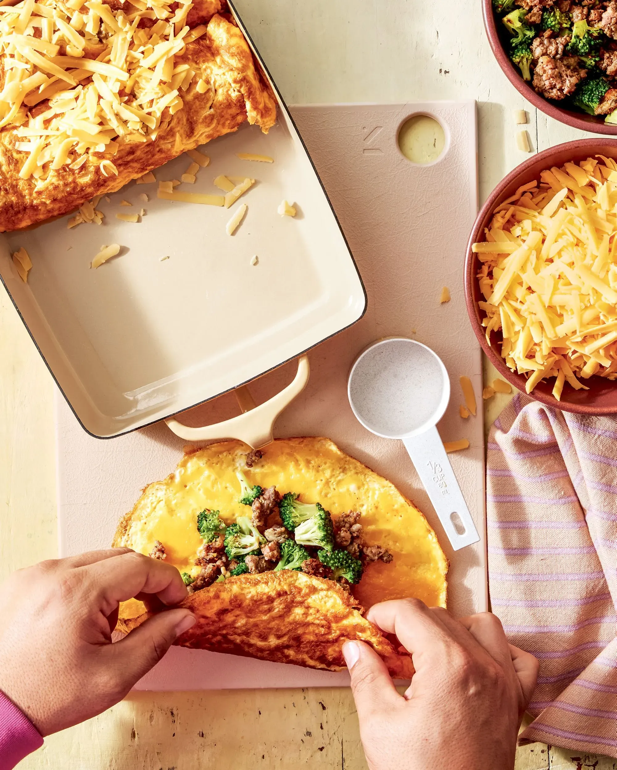 عجة دان بيلوسي المحشوة بالبروكلي والسجق، تُعد في طبق خزفي، مع جبن شيدر مبشور، وتظهر يد تُحضر العجة.