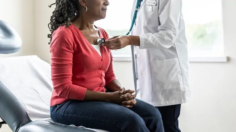 امرأة تجلس في عيادة طبية، بينما يقوم طبيب بفحص قلبها باستخدام سماعة طبية، مما يعكس أهمية تقييم صحة القلب قبل الجراحة.