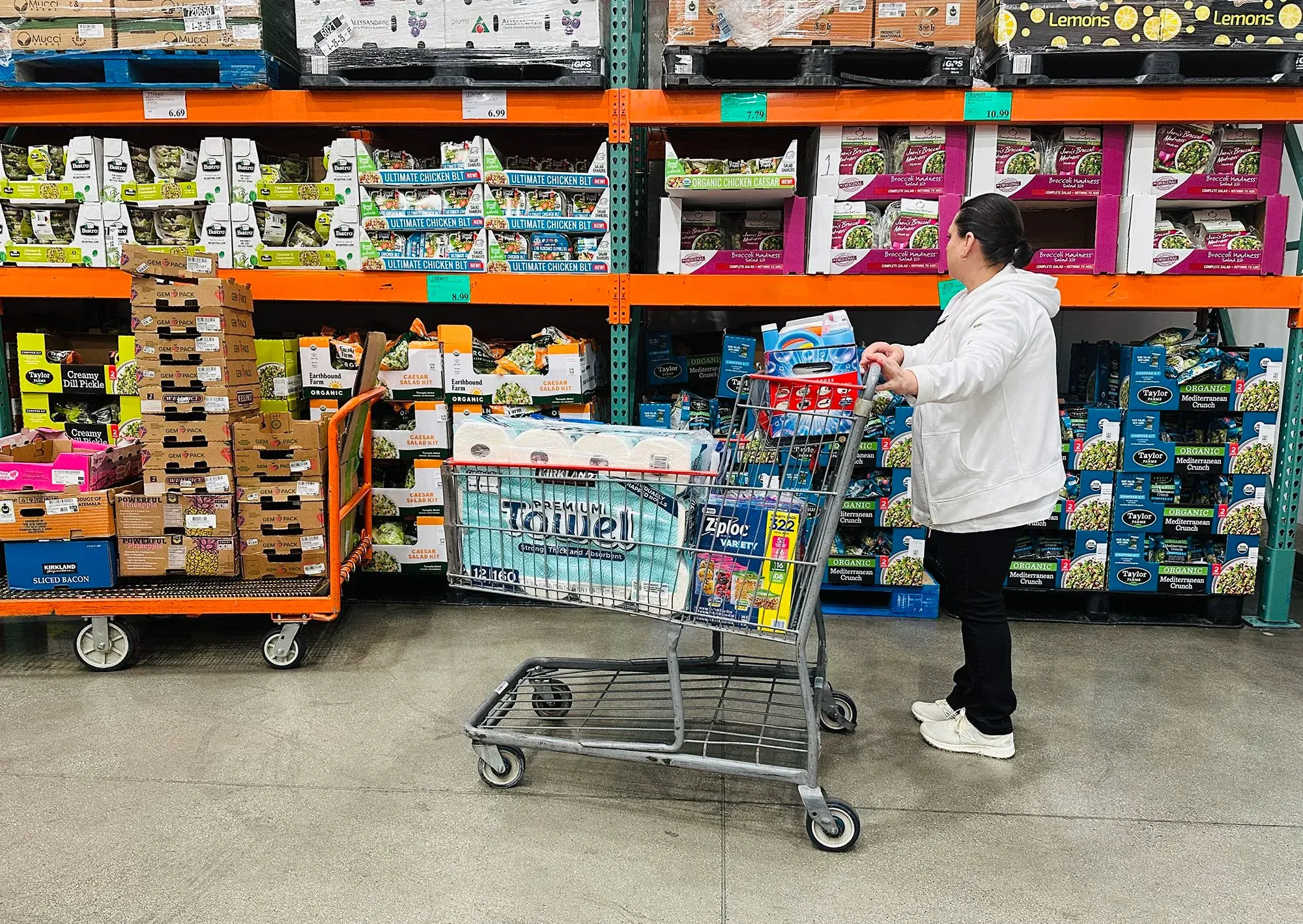 امرأة تتسوق في متجر كوستكو، حيث تظهر الأرفف المليئة بالمنتجات الغذائية ومتنوعة، مما يعكس تنوع العرض في المتجر.