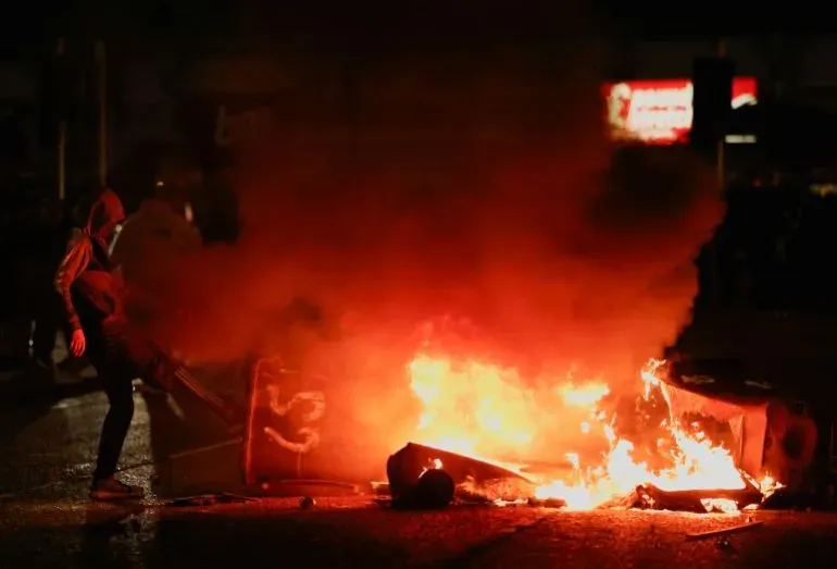 شخص ملثم يشعل النيران في حطام وسط سحب الدخان خلال اشتباكات عنيفة في أيرلندا الشمالية، تعبيرًا عن الاحتجاجات المناهضة للهجرة.