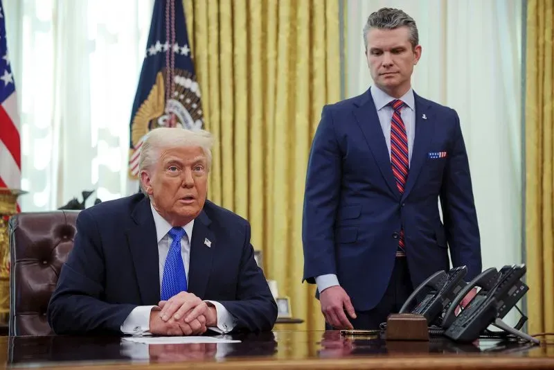 ترامب يجلس خلف مكتبه في المكتب البيضاوي بينما يقف هيغسيث خلفه، مما يعكس العلاقة بينهما في سياق الأمن القومي.